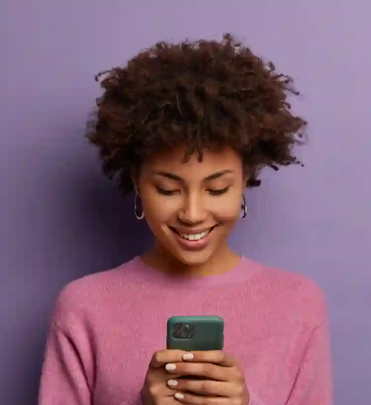 A young woman with curly hair in a pink sweater looks down at her smartphone and smiles against a solid purple background.