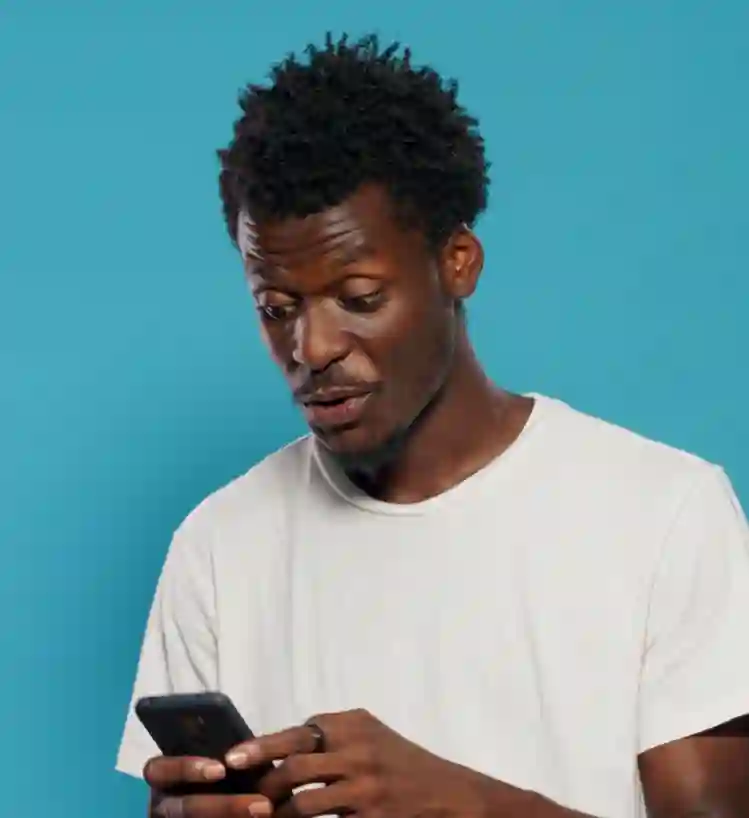 A man in a white t-shirt looking surprised while checking his smartphone against a blue background.
