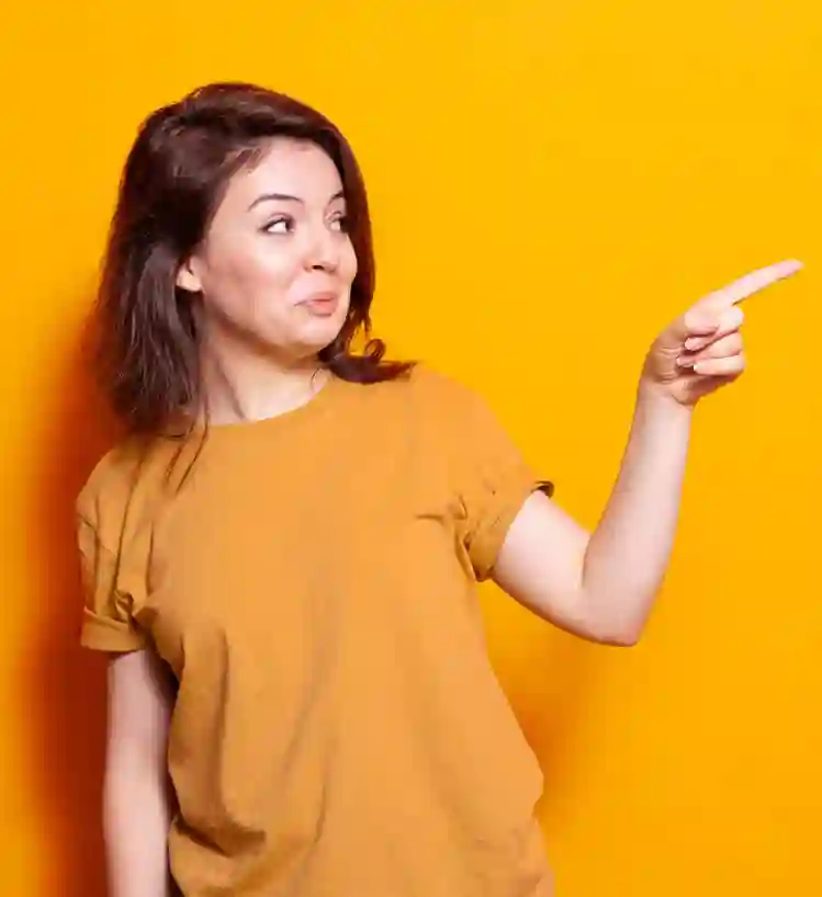 A young woman wearing an orange shirt stands in front of a bright orange background, looking and pointing to the right with her index finger.